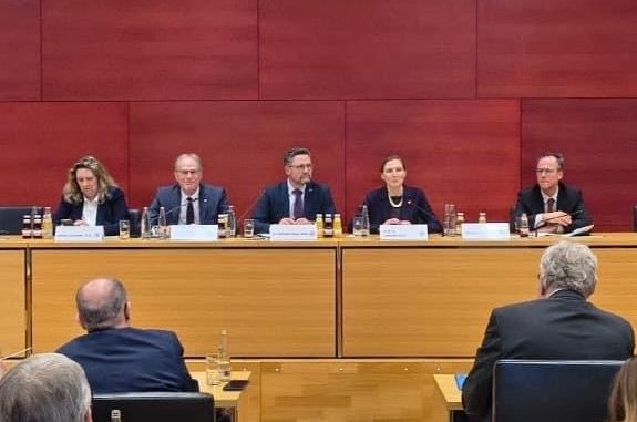 Podiumsdiskussion mit fünf Personen, die an einem Tisch sitzen. Roter Hintergrund.
