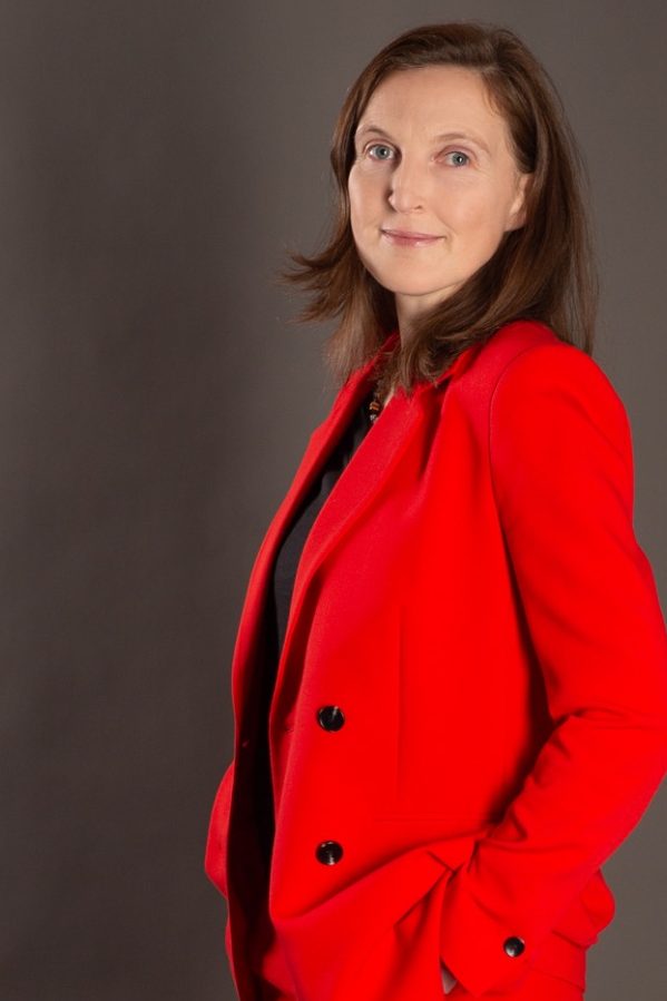 Gerlinde Groitl Porträtfoto 1 Gerlinde Groitl in rotem Blazer, seitlich blickend, lächelt freundlich vor grauem Hintergrund.