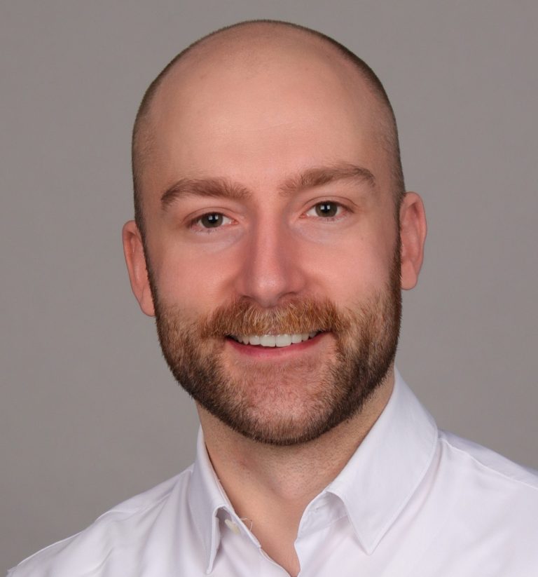 Porträt eines Mannes mit Bart, lächelnd, in einem weißen Hemd.