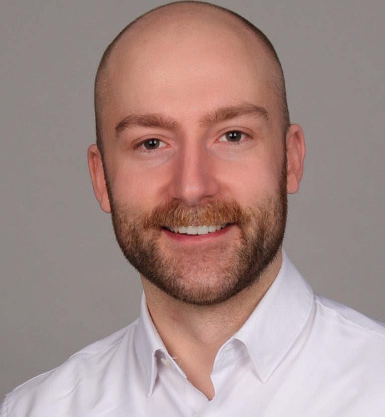 Fabian Frenger M.A. Lächelnder Mann mit Bart, in einem weißen Hemd.