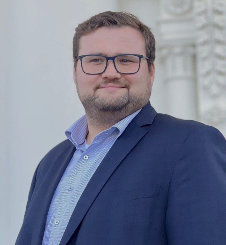 Christoph Götz B.A. Mann mit Brille, lächelt, trägt Anzug und steht vor einer weißen, dekorativen Wand.