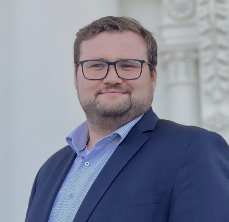 Mann mit Brille in Anzug und Hemd, lächelt vor einem hellen Hintergrund.