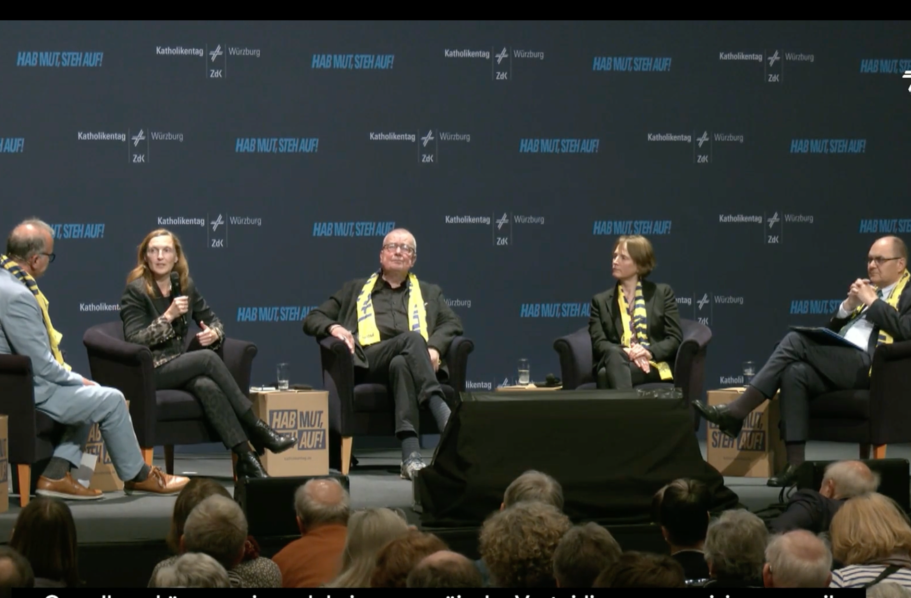 Konferenz mit mehreren Sprechern an einem Tisch vor einer Logo-Wand.