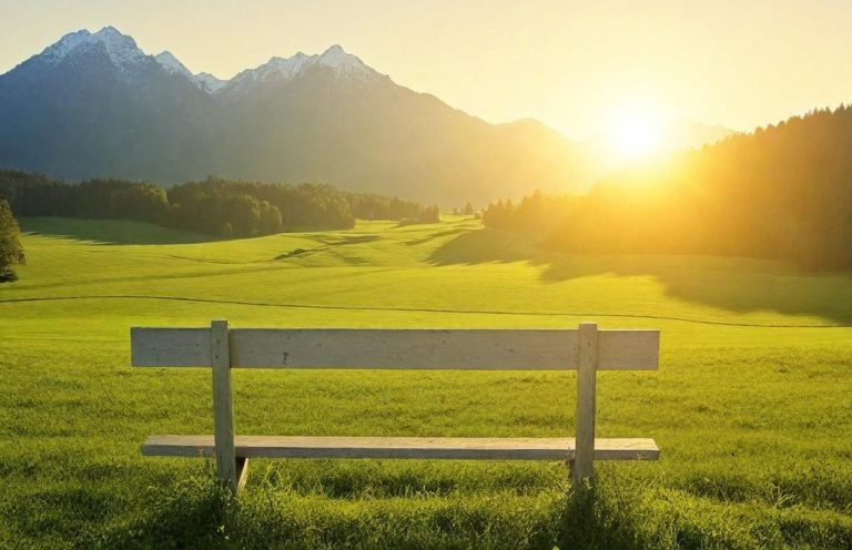 Eine Holzbank auf einer Wiese mit Bergen im Hintergrund und Sonnenaufgang.